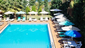 A swimming pool with tiled edges, surrounded by white lounge chairs and umbrellas, set in a landscaped garden with trees and a lawn. - Kalithea Apartments (Apokoronas)