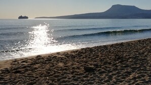 A beach with a ship in the distance, a mountain, and the sun reflecting on the water. - Kalithea Apartments (Apokoronas)