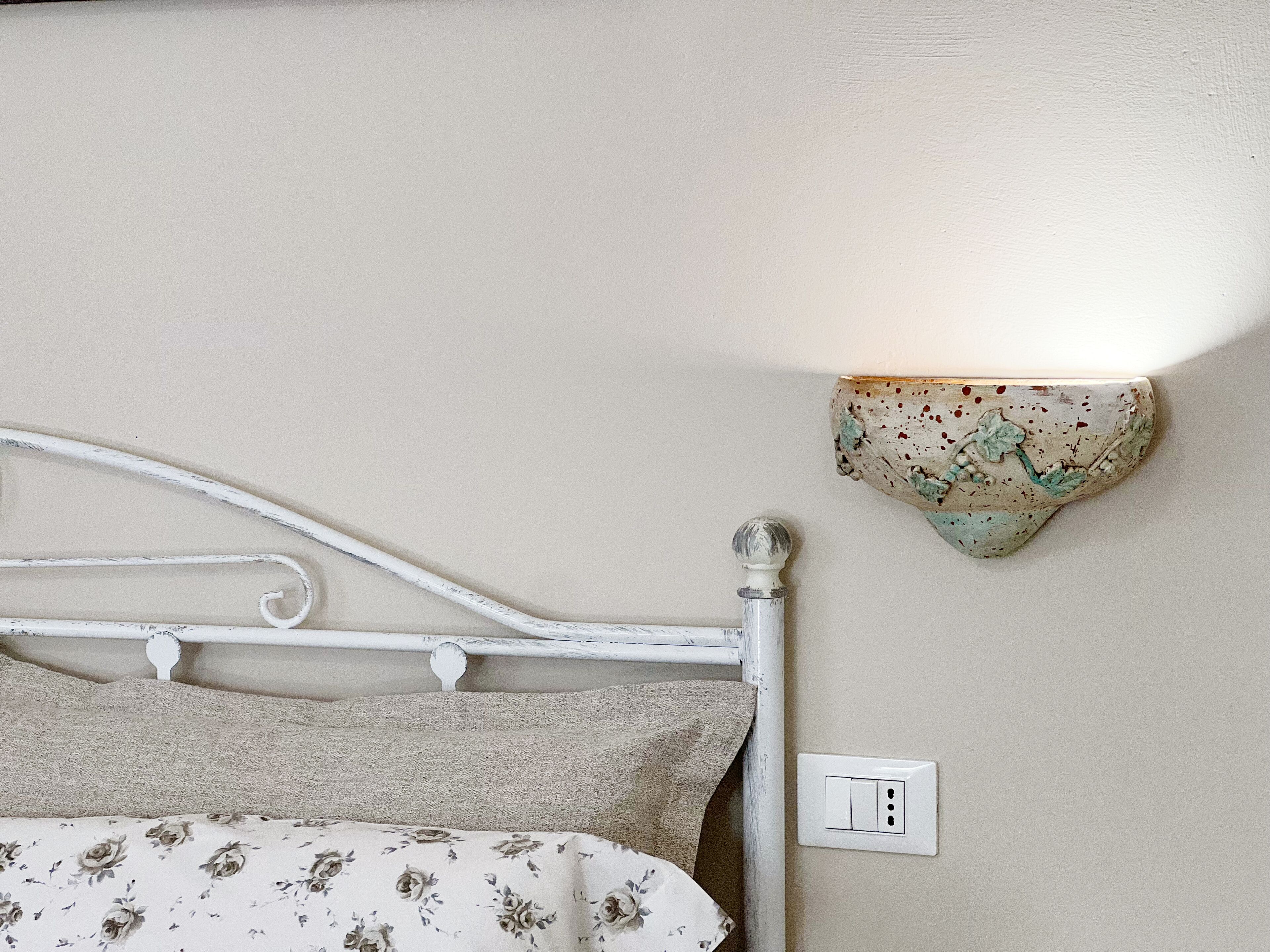 Una cama con una colcha estampada con motivos florales y un cabecero metálico blanco. Se aprecia una lámpara de pared con diseño decorativo.