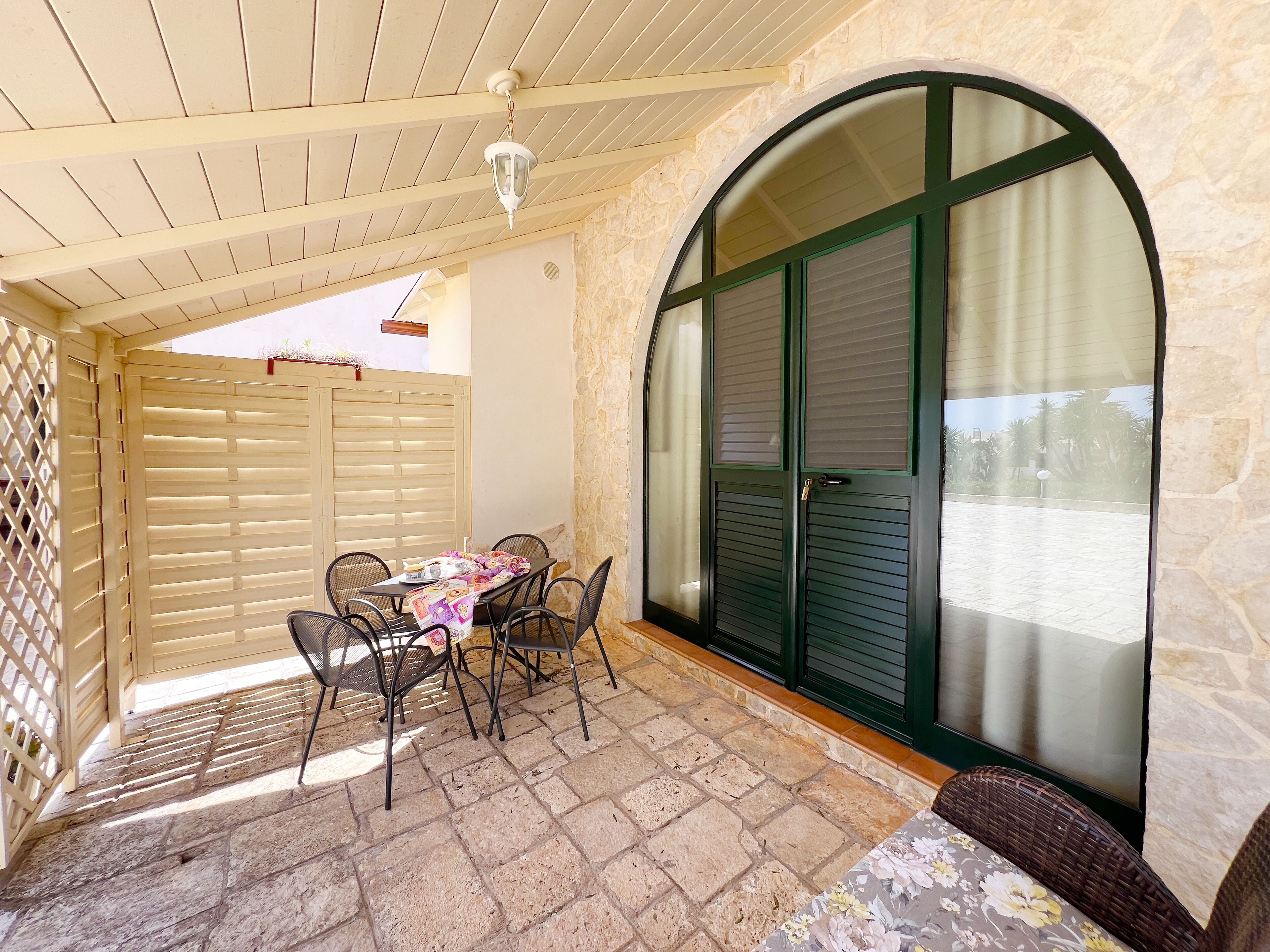 A covered patio with a table and chairs, stone flooring, and a view through a large arched window.