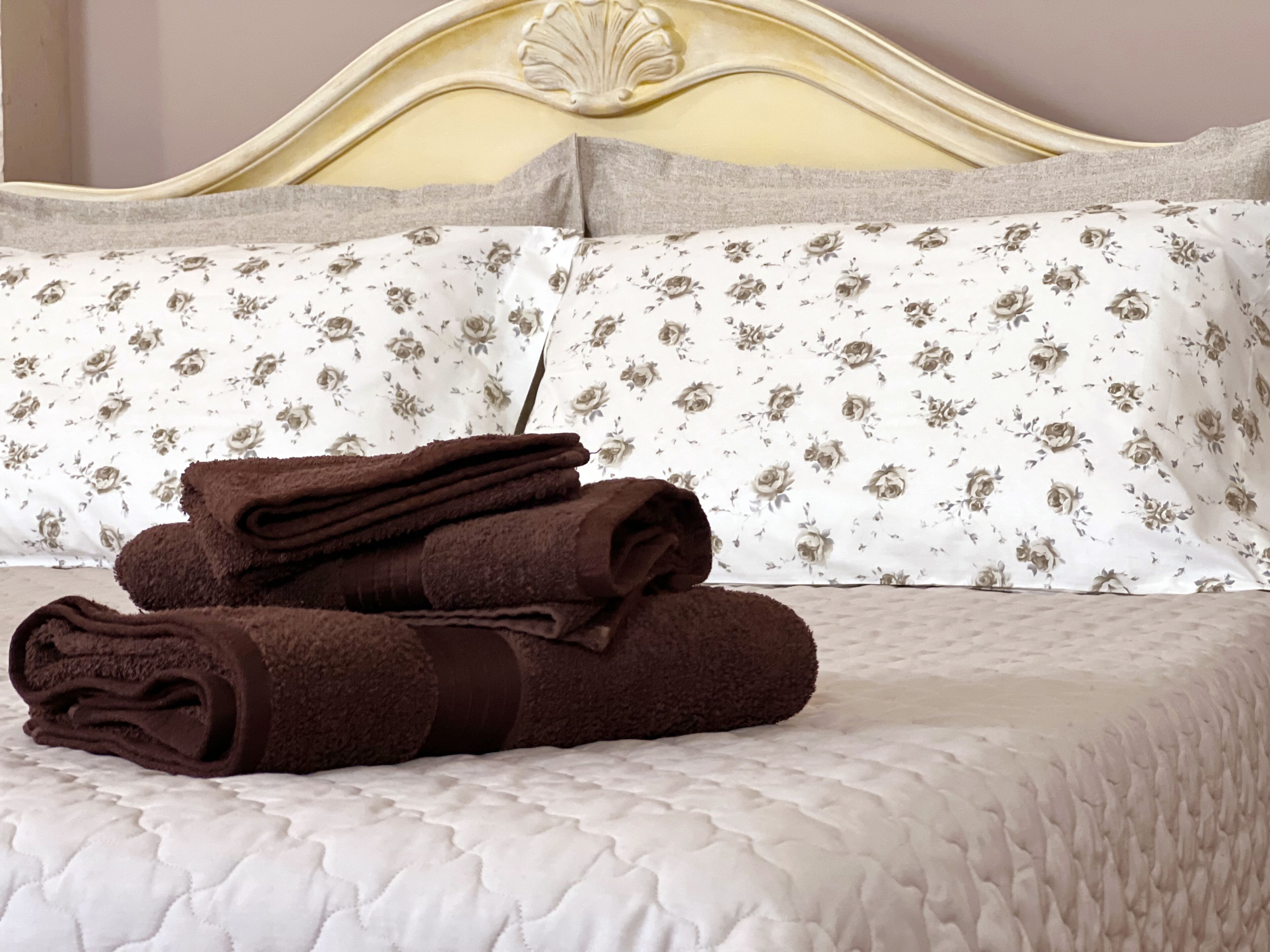 Un lit bien fait, avec une couette blanche matelassée, un coussin à motifs floraux et une pile de serviettes marron pliées.