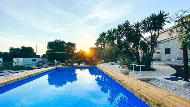 A swimming pool with a wooden deck and lounge chairs, surrounded by palm trees and a building with a stone facade. - La Maison One B&B (Carovigno)