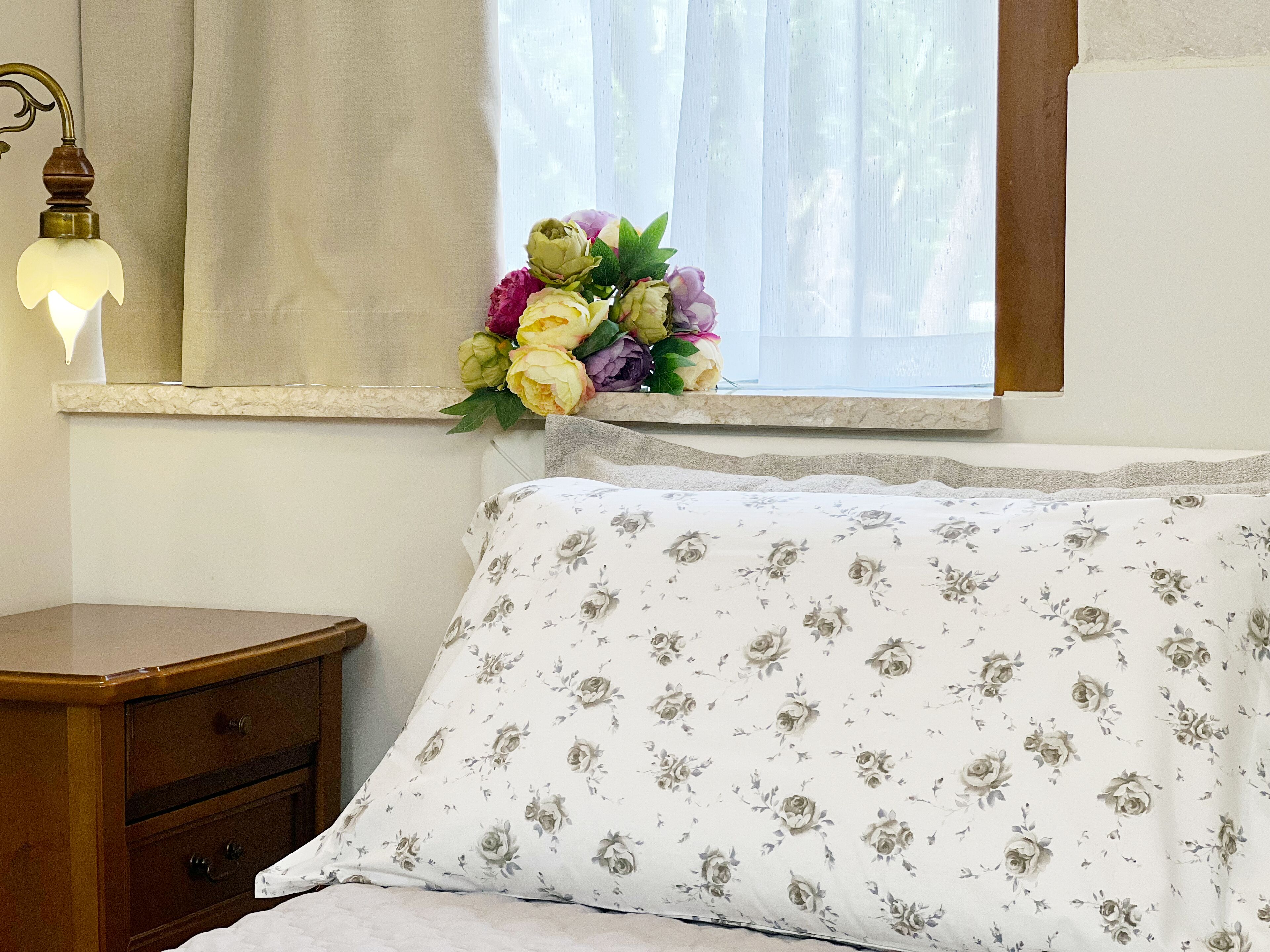 A bedroom with a bed, a nightstand, a lamp, and a window with a floral arrangement.