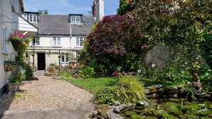 A quaint cottage with a gravel driveway leading to a wooden door, surrounded by lush gardens and a small pond with lily pads. - Ammonite Lodge Guest House (Chard)