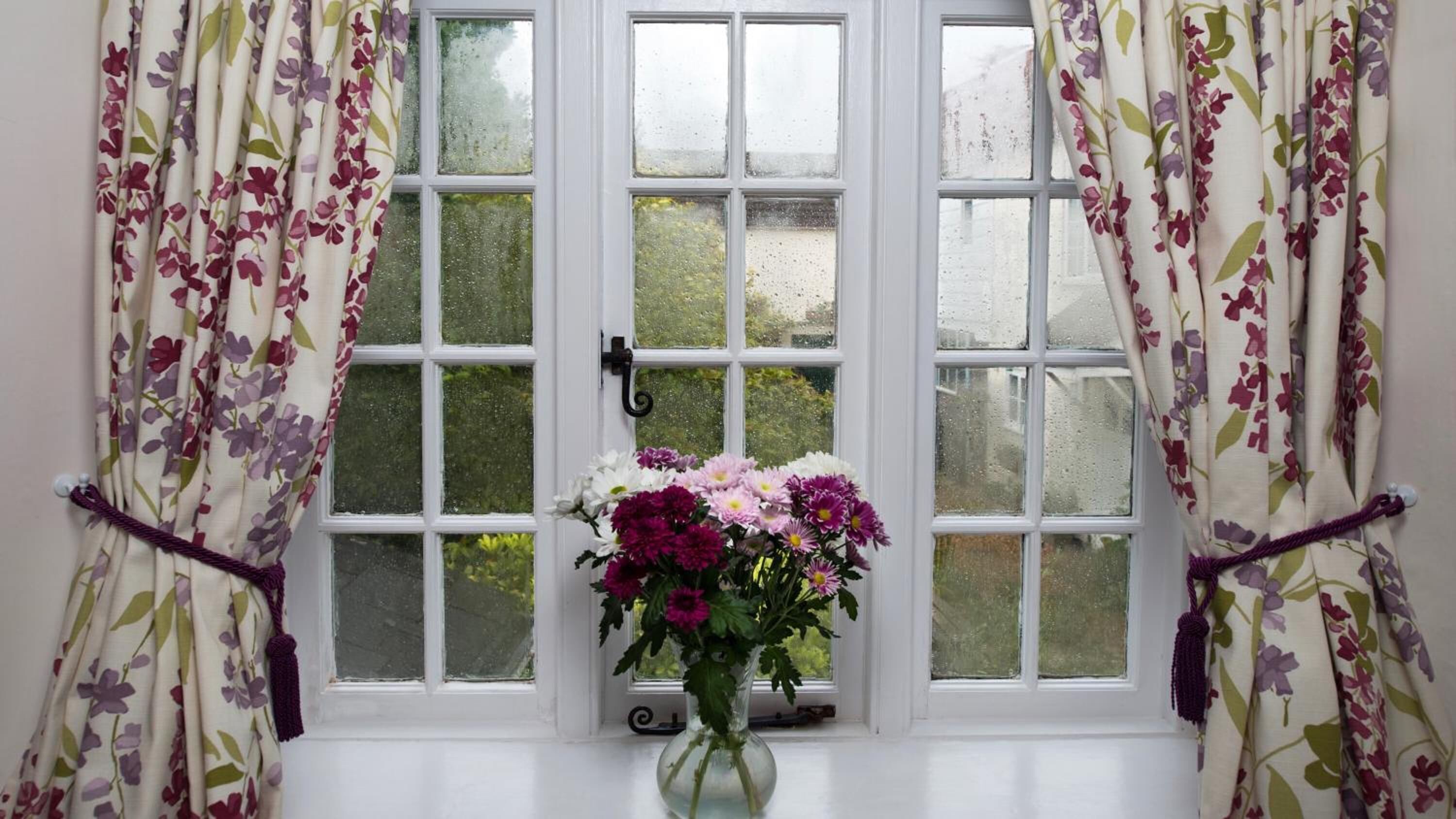 Una ventana con cortinas florales, vistas claras a la vegetación exterior y un florero con flores en el alféizar.