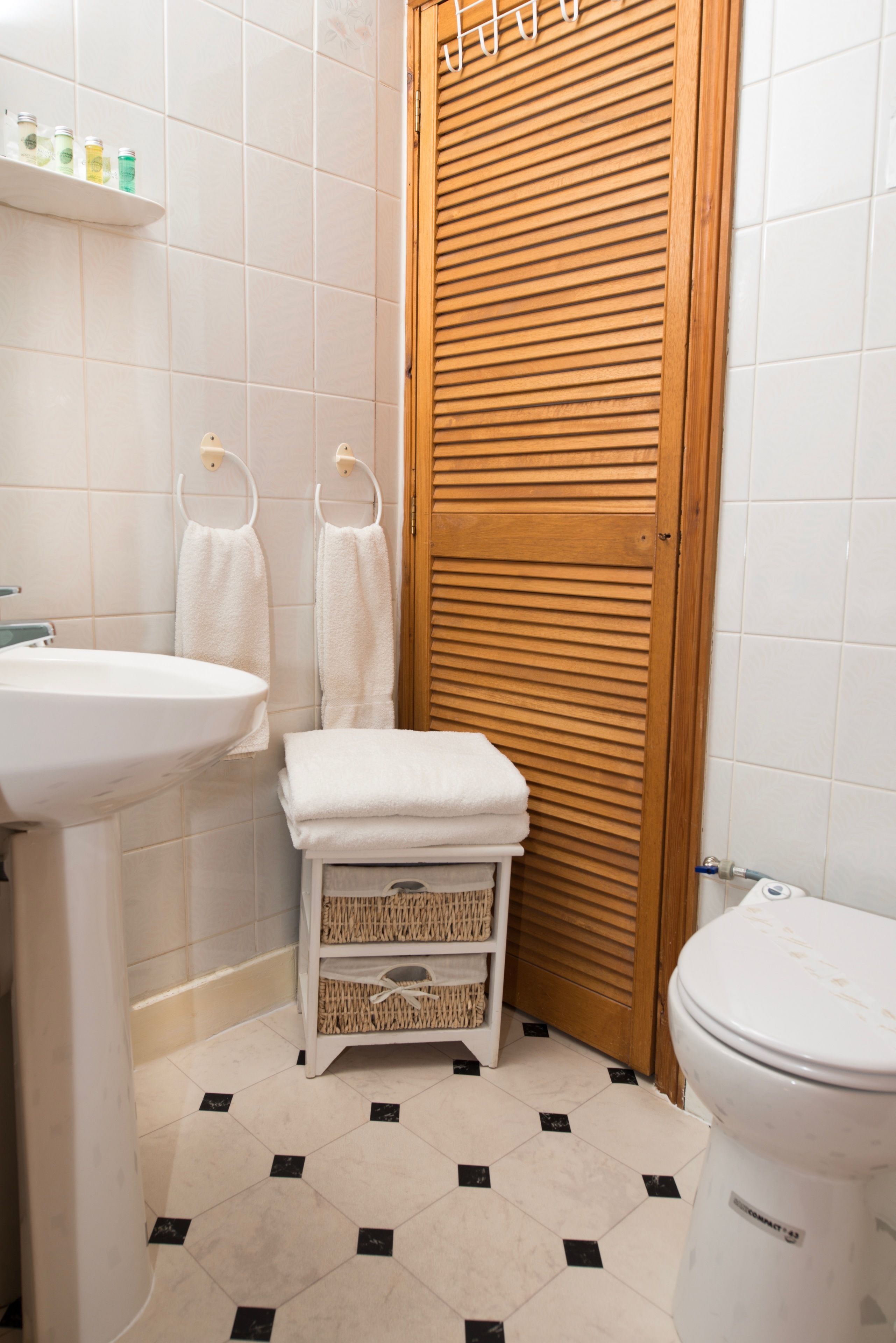 Un baño con lavamanos, inodoro y puerta de madera. Hay toallas colgadas en ganchos y una cesta en el suelo.