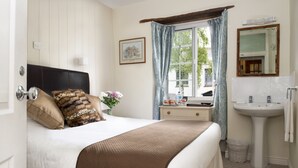 A bedroom with a bed, a bedside table, a window with curtains, a bathroom with a sink and mirror. - Ammonite Lodge Guest House (Chard)