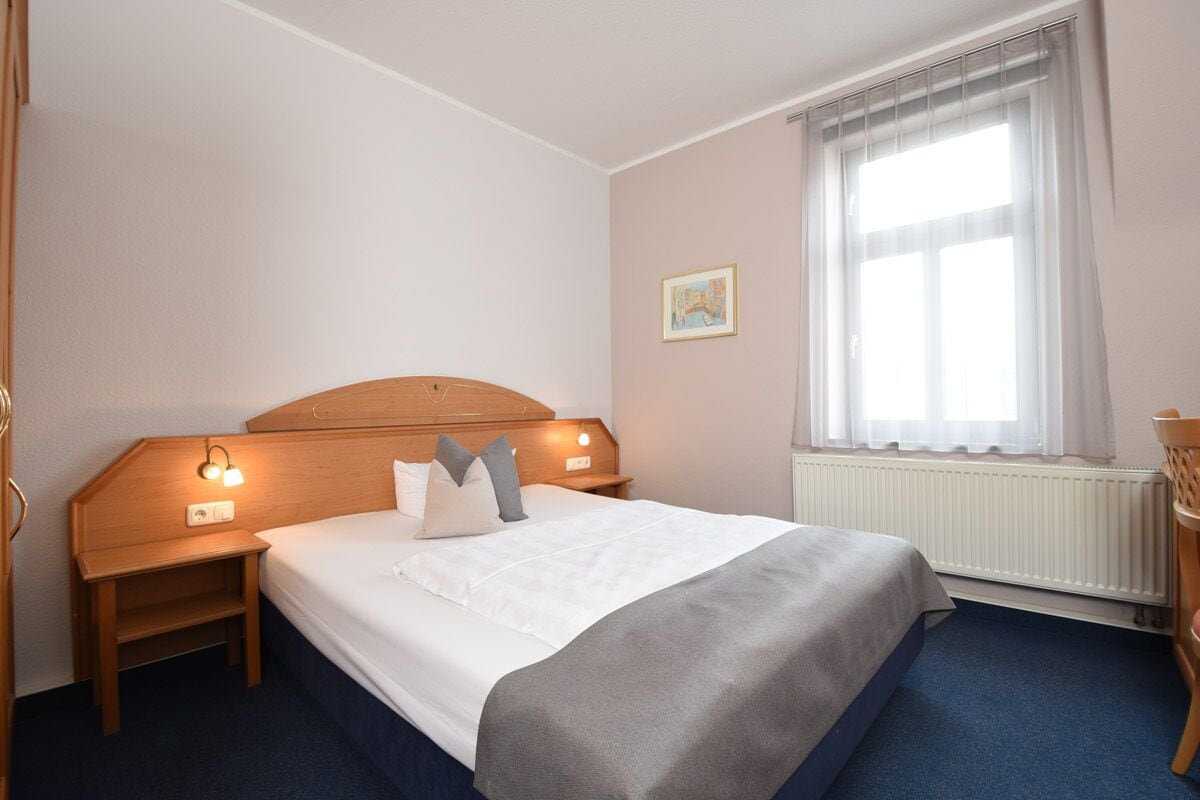 A hotel room with a single bed, a nightstand, a wall-mounted lamp, a window with sheer curtains, and a radiator.