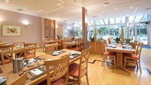 Un comedor amplio con varias mesas y sillas de madera, ventanas grandes que ofrecen vista a la vegetación y un diseño moderno del techo.