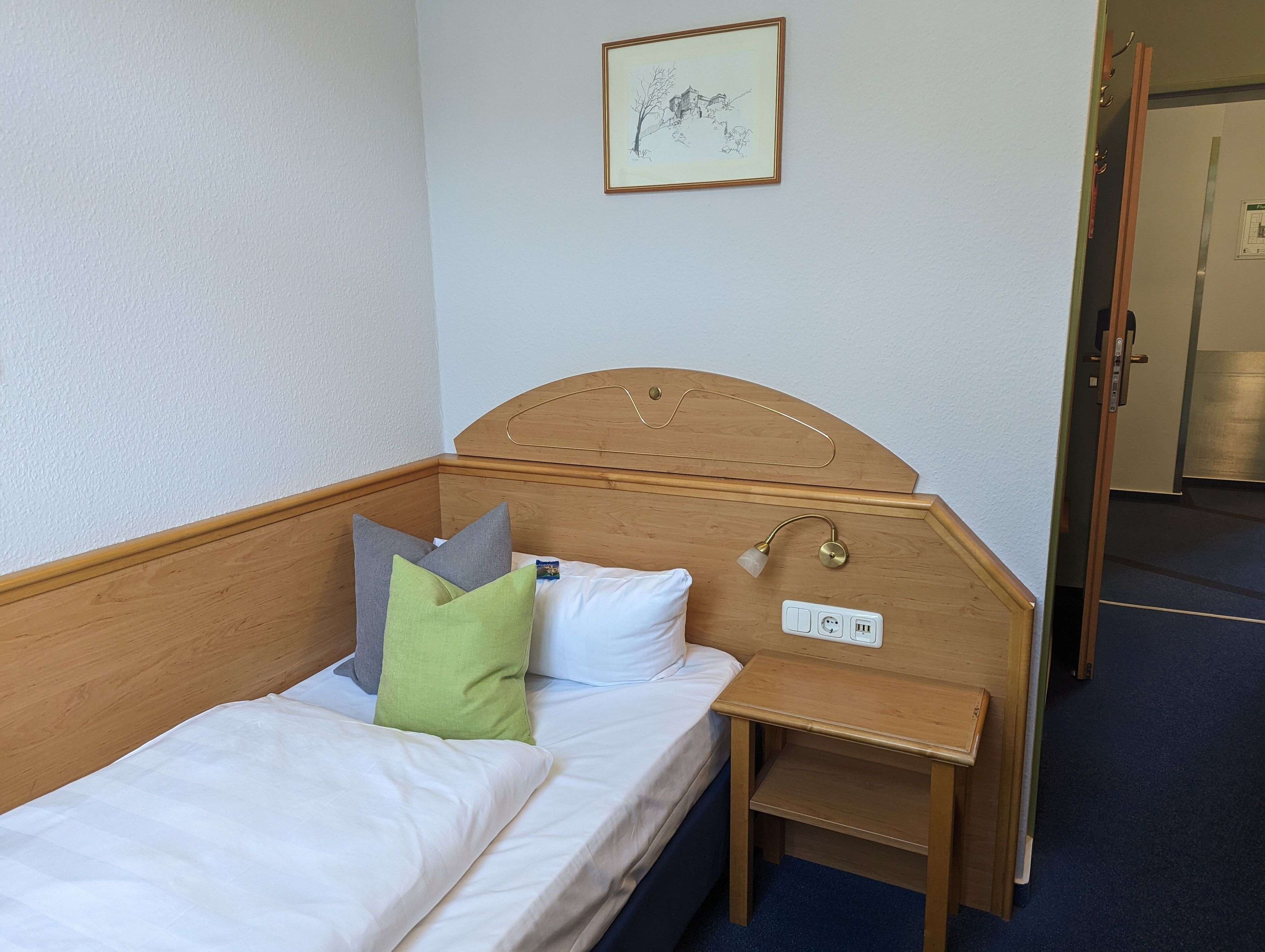 Un lit simple avec une tête de lit en bois, une table de chevet avec une lampe et une prise de courant.