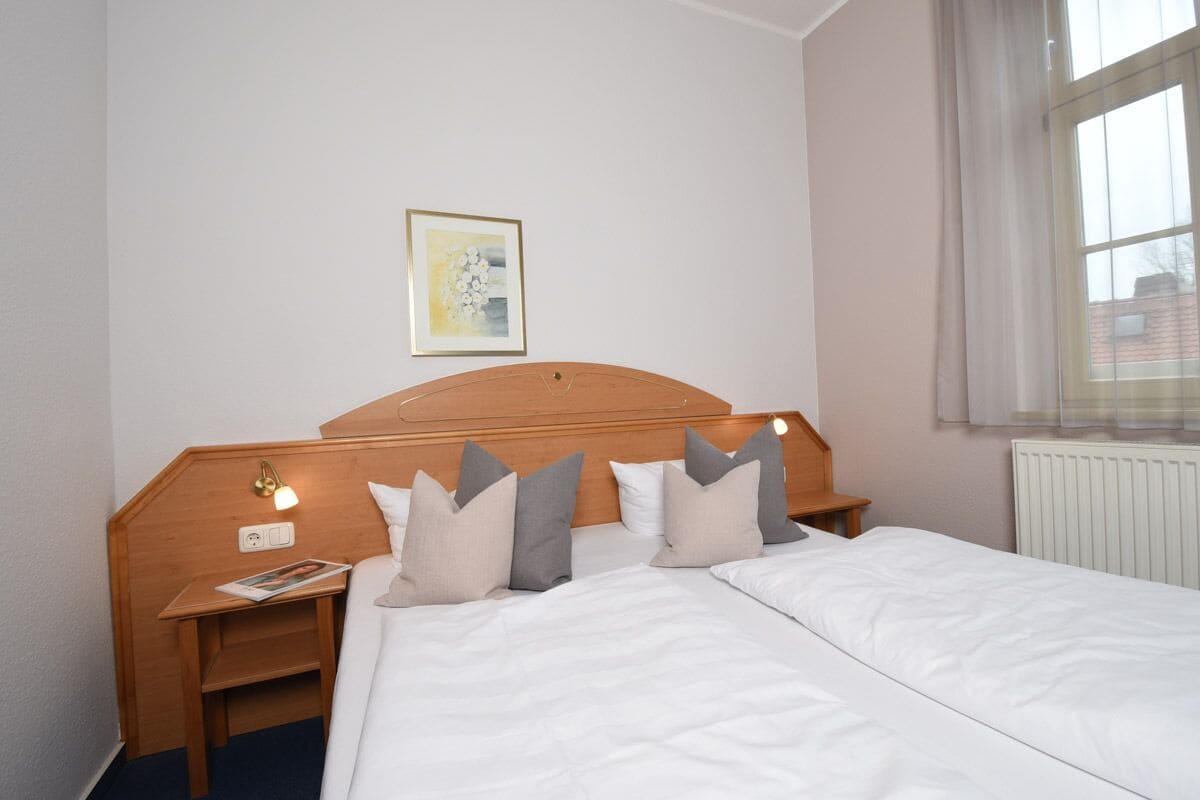 Habitación de hotel con cama doble, cabecero de madera, mesitas de noche con lámparas, un cuadro enmarcado en la pared y una ventana con cortinas.