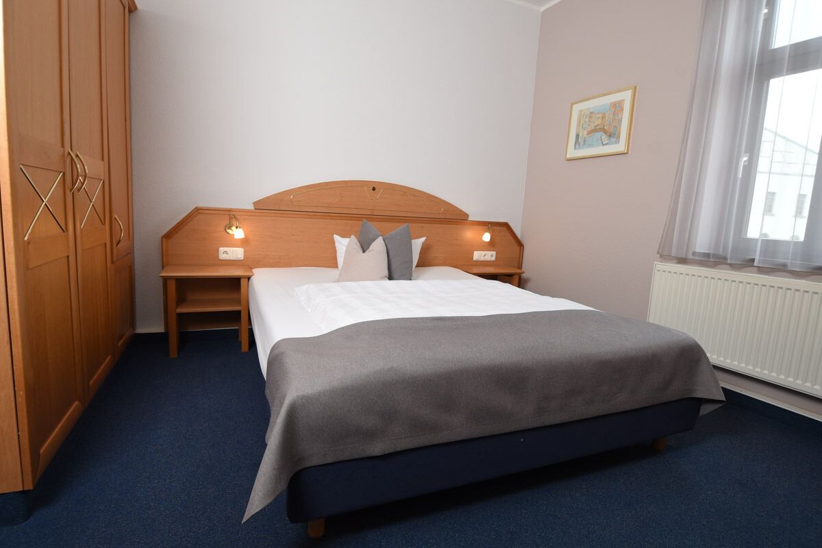 A bedroom with a bed, wooden wardrobe, bedside lamps, a radiator, and a window with curtains.