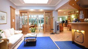 Un vestíbulo de hotel con mostrador de recepción, una zona de estar con sofá y mesa de centro, y vistas al área de comedor exterior.