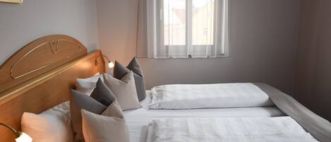Un dormitorio con cabecera de madera, dos camas con ropa blanca y una ventana con cortinas de gasa.