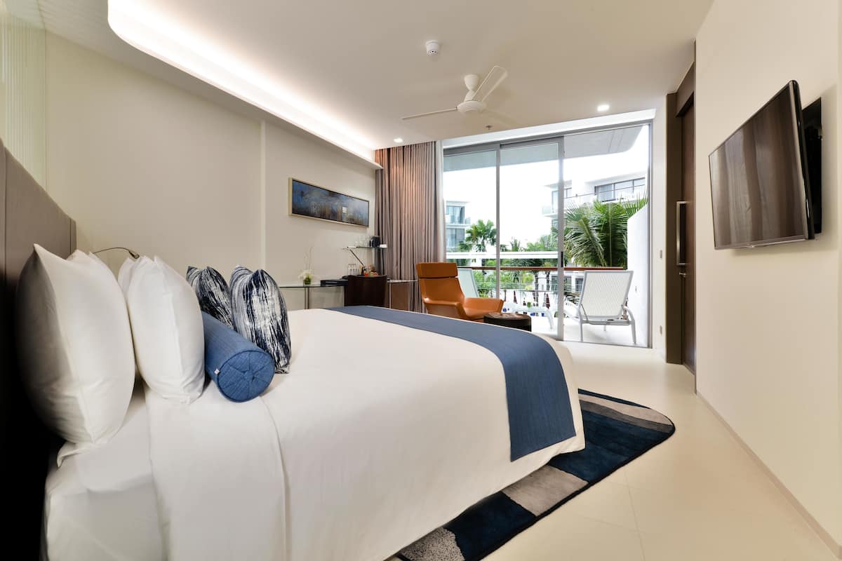 A modern hotel room with a large bed, a flat-screen TV, a balcony with a view of palm trees, and a ceiling fan.