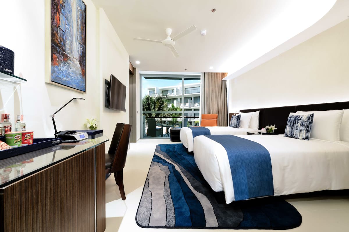 A modern hotel room with a large bed, a desk with a computer, a flat-screen TV, and a large window with a view of buildings and greenery.