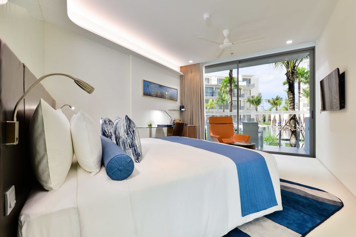 A modern hotel room with a large bed, a desk, a chair, and a balcony with a view of palm trees and buildings.