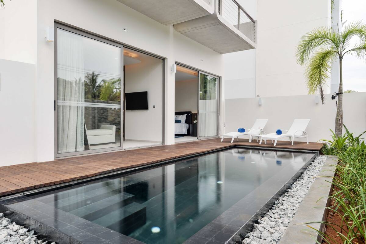 A modern outdoor swimming pool with a wooden deck, adjacent to a white building with large glass doors and a flat roof.