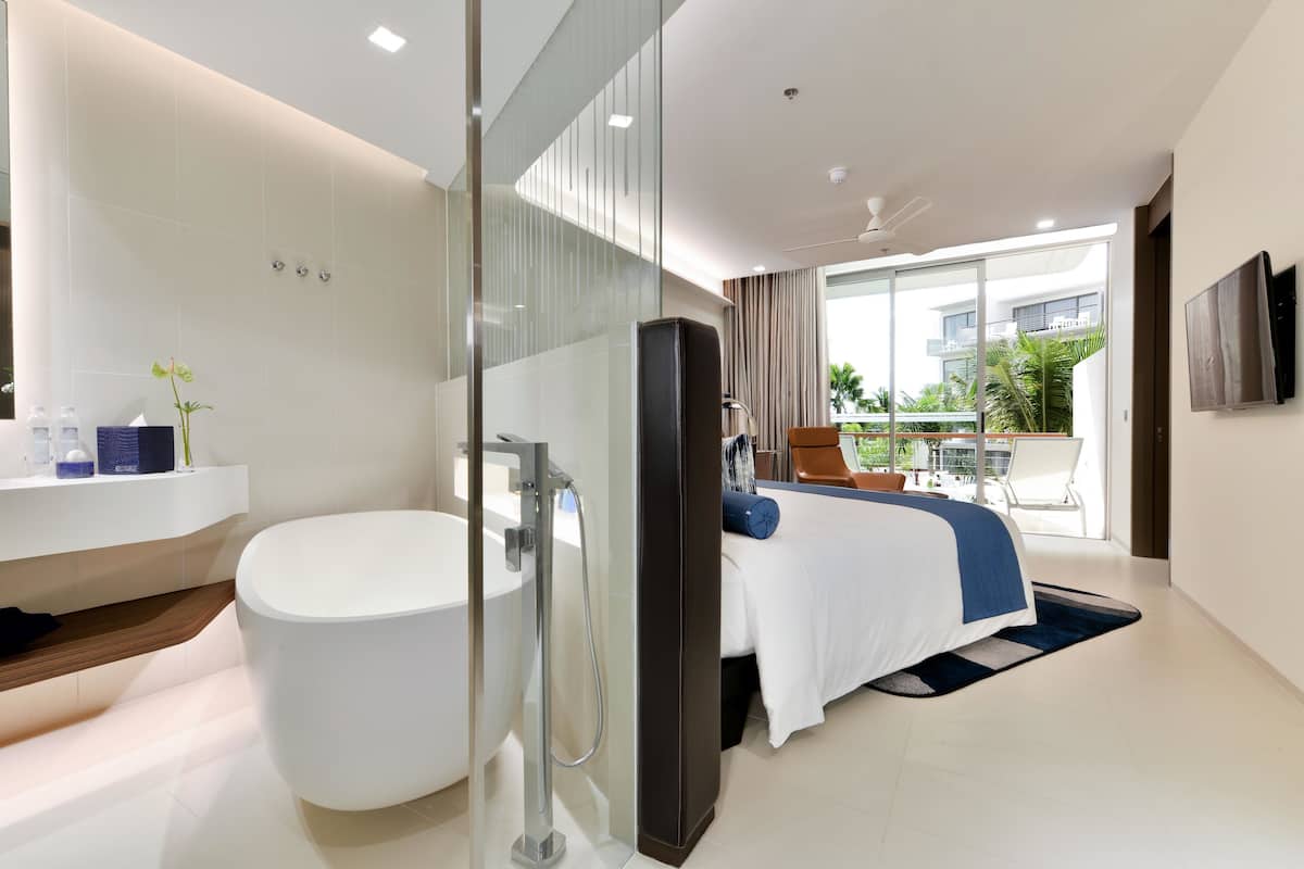 A modern hotel room with a large bathtub, a bed with a blue accent, a glass shower enclosure, and a balcony with a view of the city.