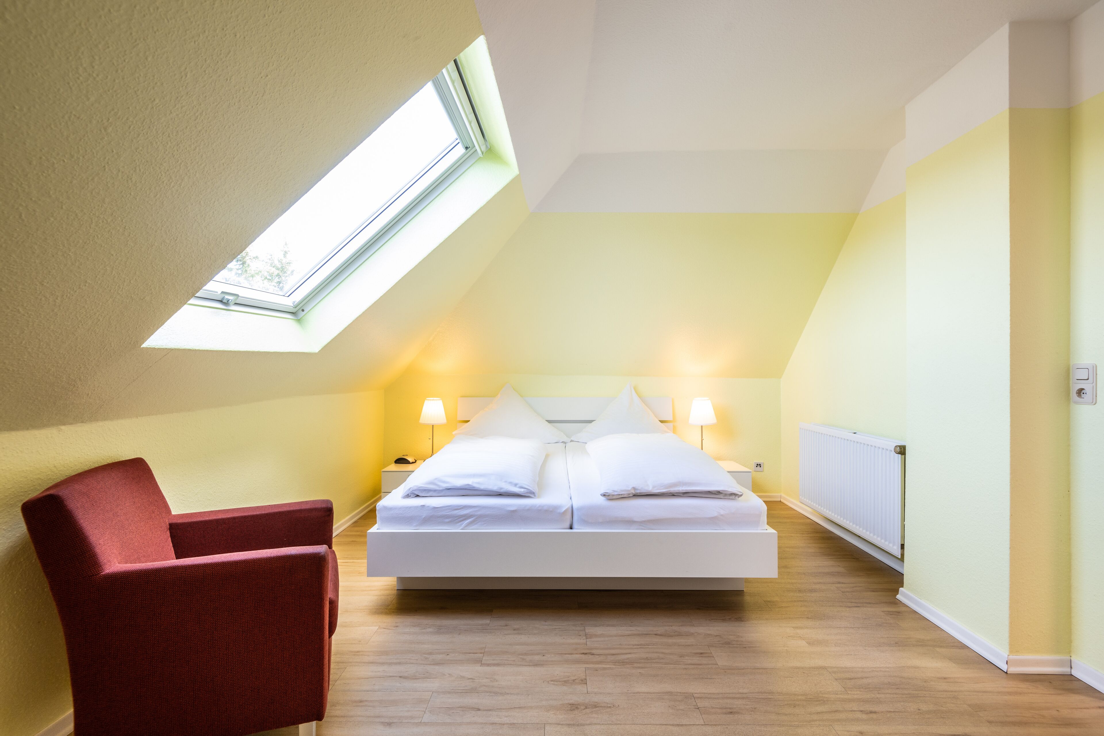 A bedroom with a skylight, a bed with white linens, two bedside lamps, a red chair, and a radiator.