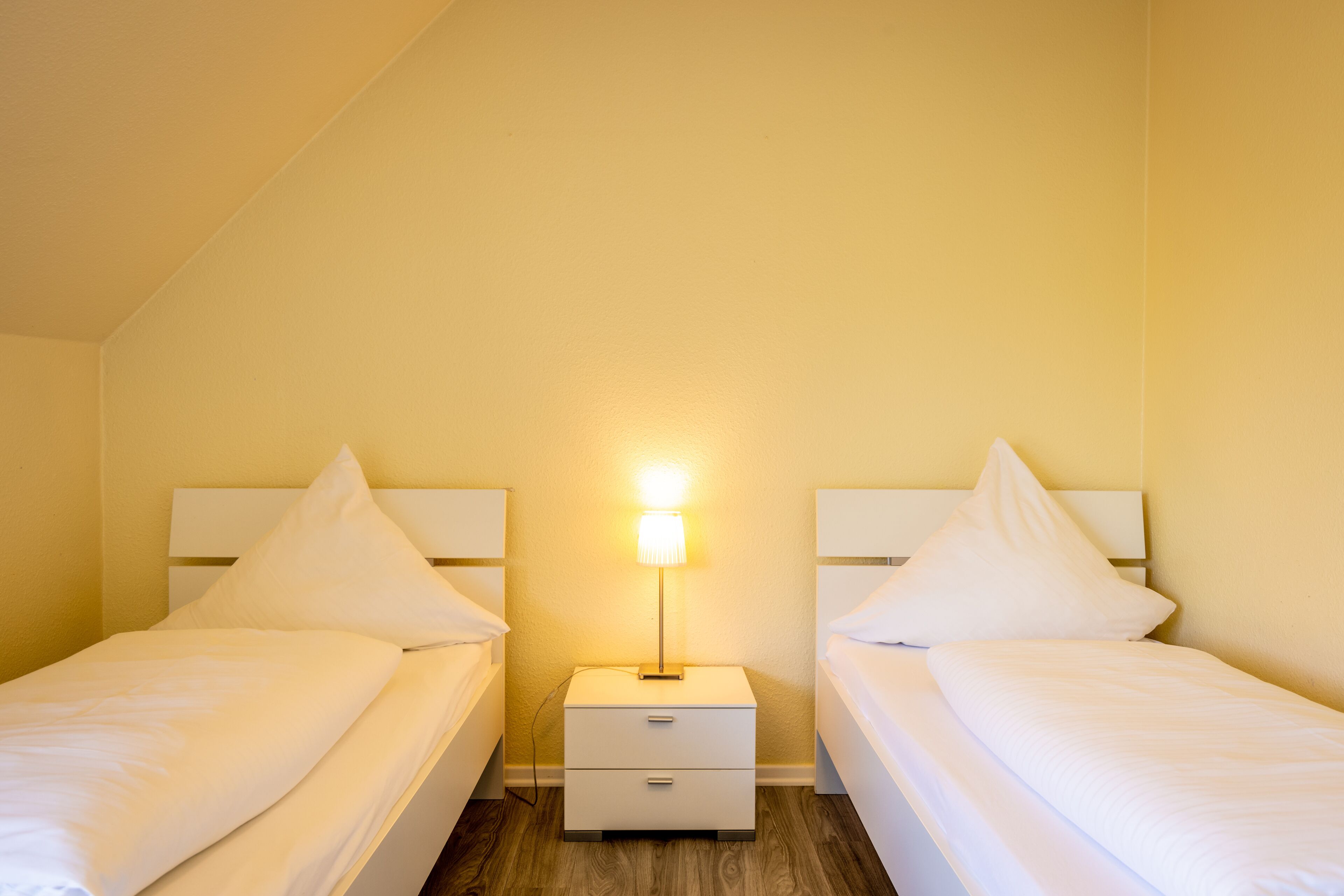 Habitación doble con dos camas, una mesita de noche con lámpara y suelo de madera.