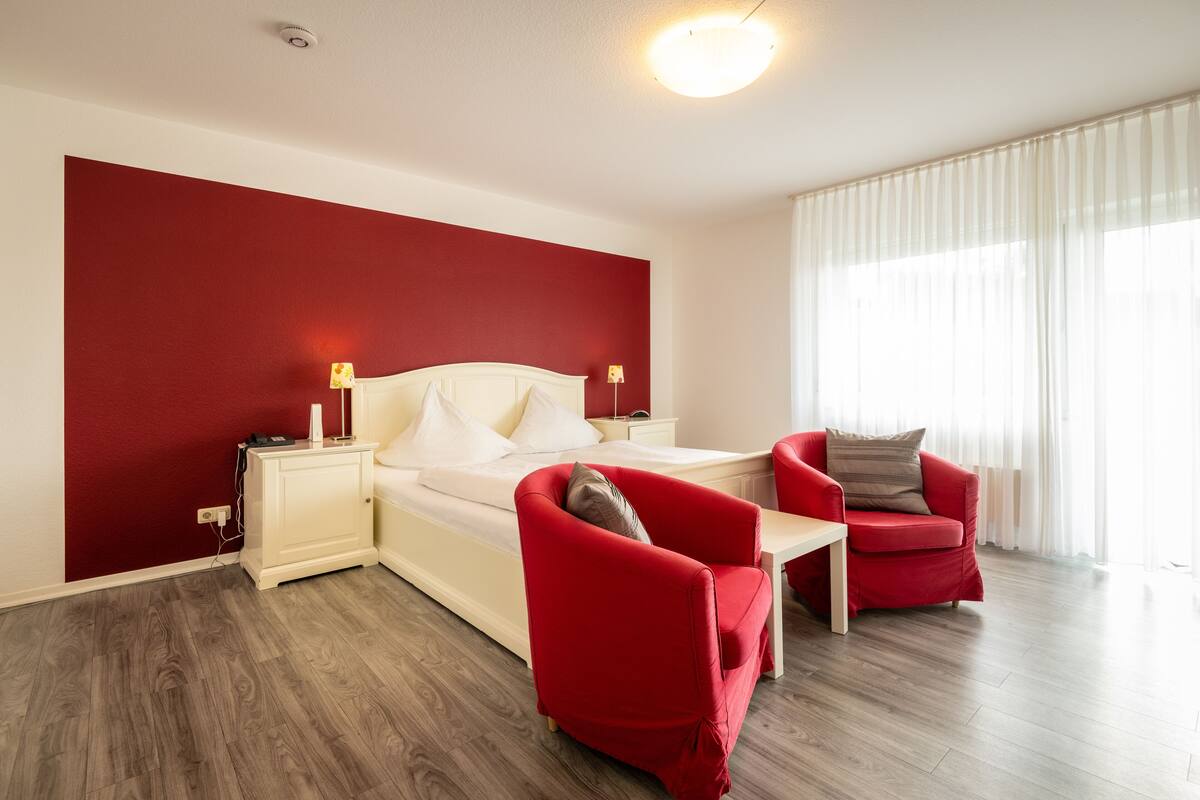 Une chambre d’hôtel moderne avec un mur d’accent rouge, un lit avec du linge de maison blanc, un fauteuil rouge, une petite table et une grande fenêtre avec des rideaux légers.