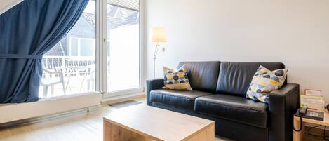 A modern living room with a black leather sofa, wooden coffee table, and a view of a balcony through a large window with blue curtains.