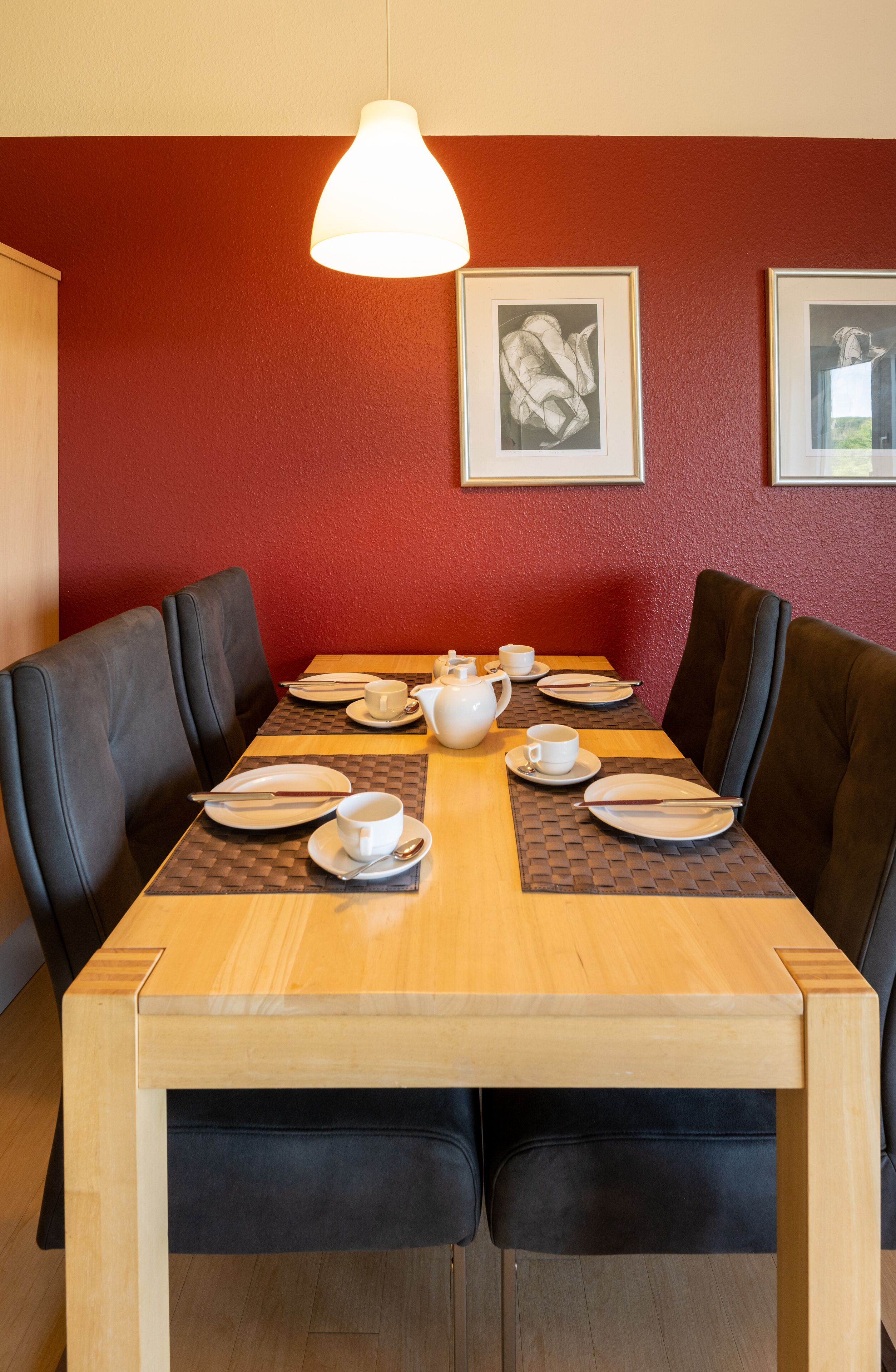 A dining area with a wooden table set for four, black upholstered chairs, a white table lamp, and two framed pictures on the wall.