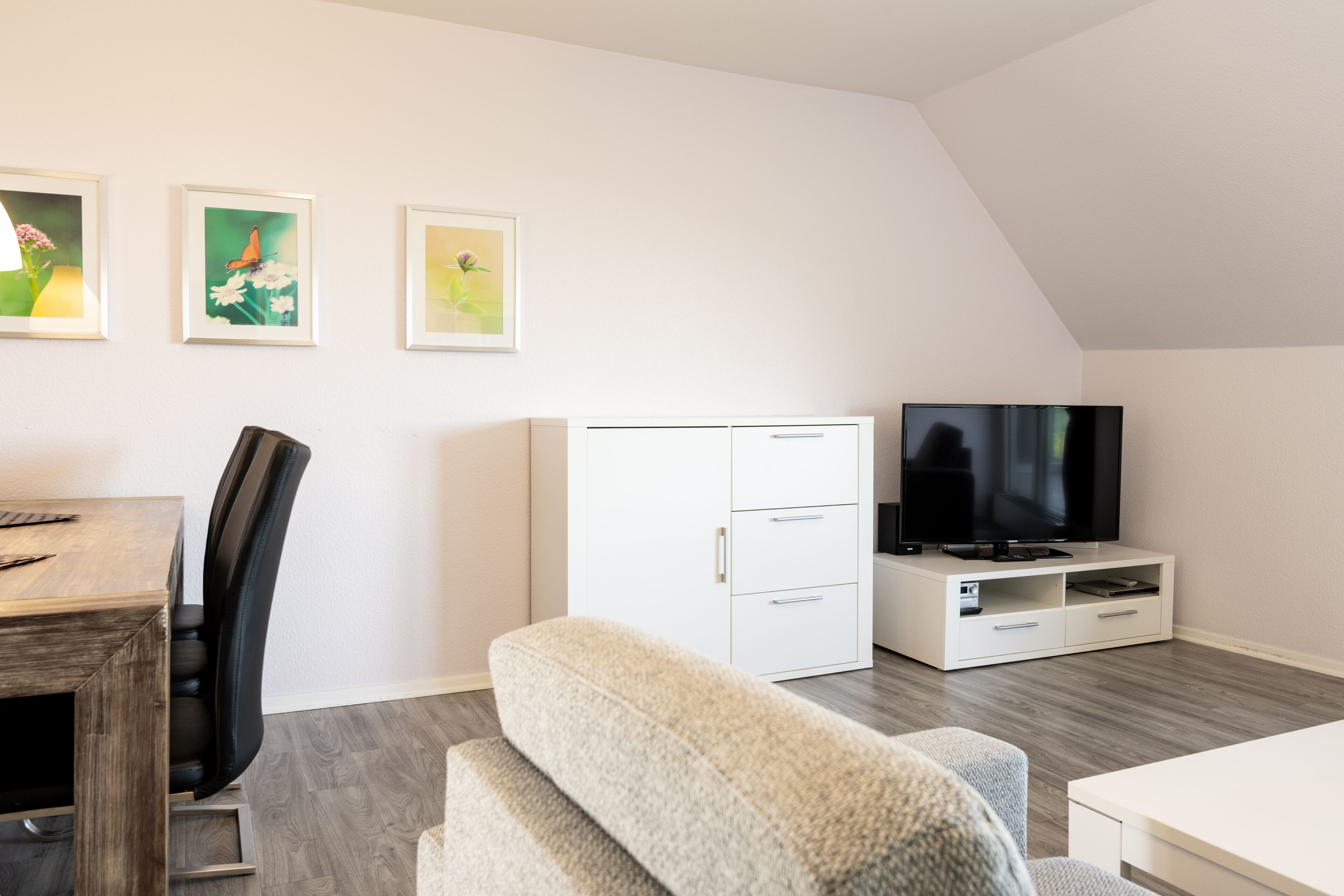 A modern living room with a wooden dining table, a sofa, a TV on a white entertainment unit, and three framed artworks on the wall.