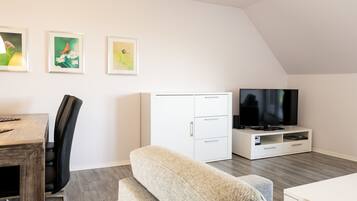 A modern living room with a wooden dining table, a sofa, a TV on a white entertainment unit, and three framed artworks on the wall.