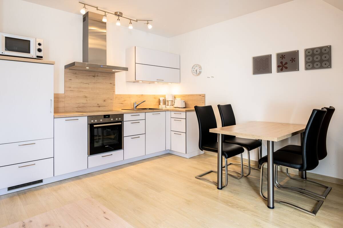 Une cuisine moderne avec des armoires blanches, un comptoir en bois, un four encastré, un four à micro-ondes et un coin repas comprenant une table et des chaises.