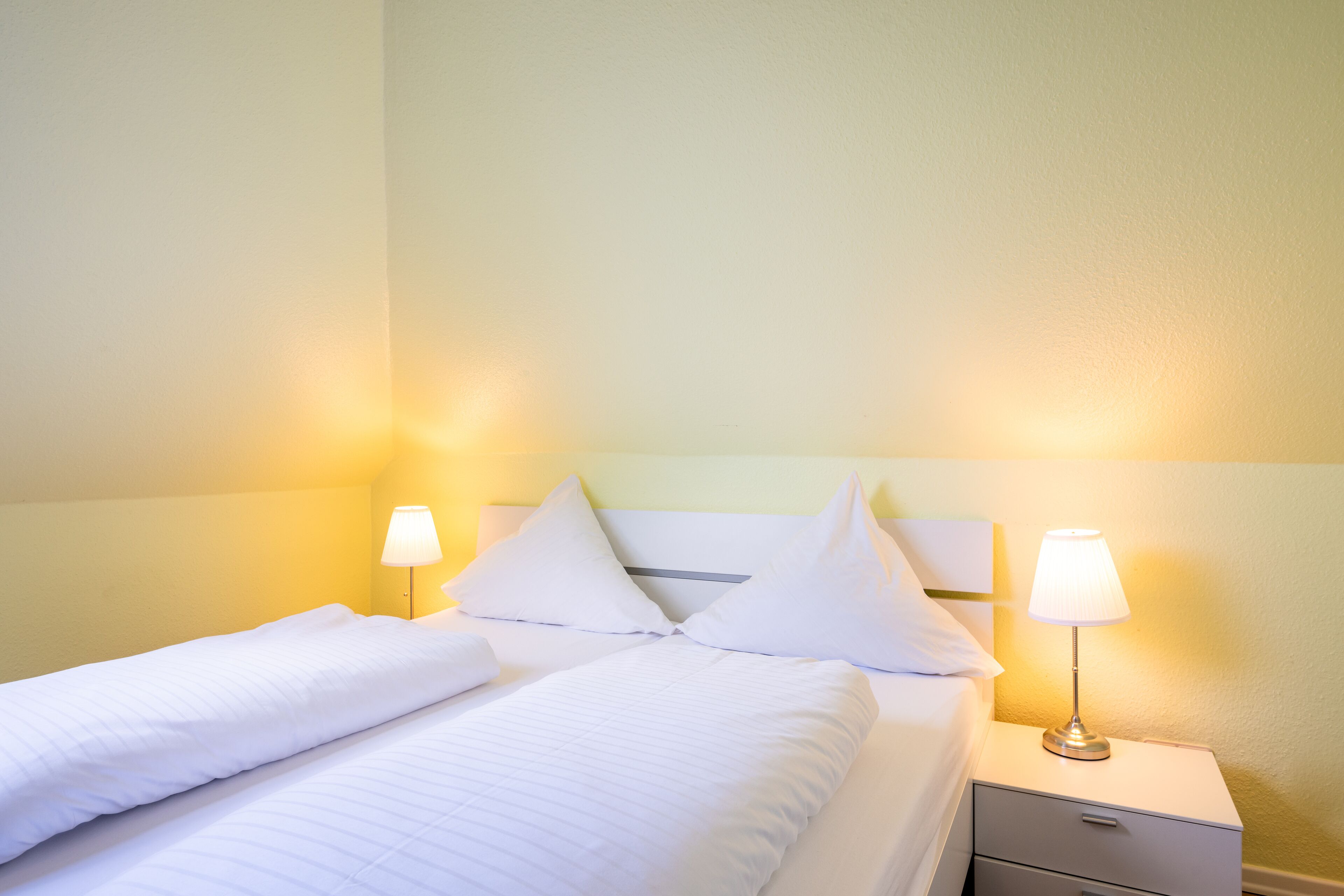 Una cama bien hecha con sábanas y almohadas blancas, una lámpara de noche y un pequeño cajón debajo de la mesita.