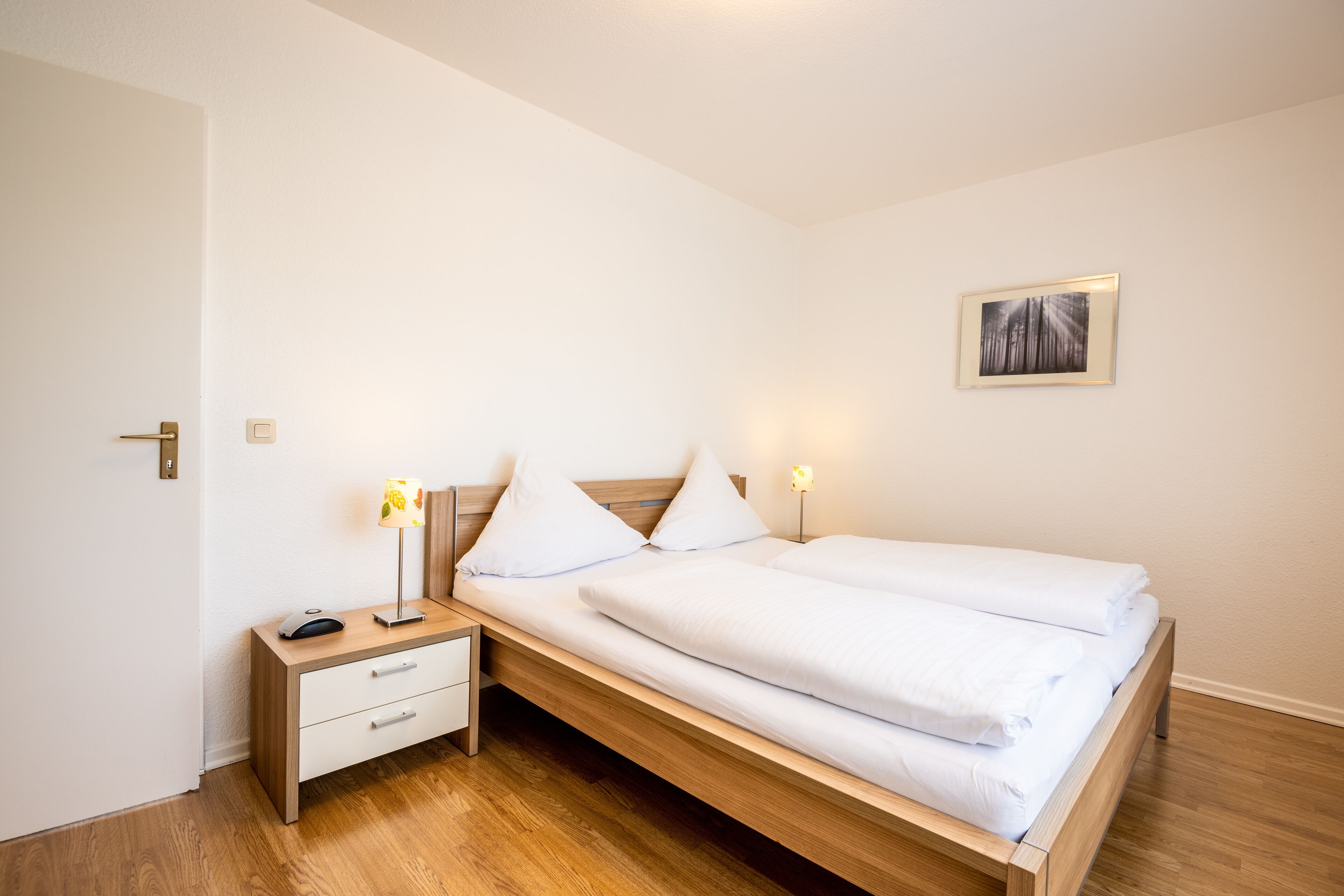 A neatly made bed with white linens, a wooden bedside table with a lamp, and a framed picture on the wall.