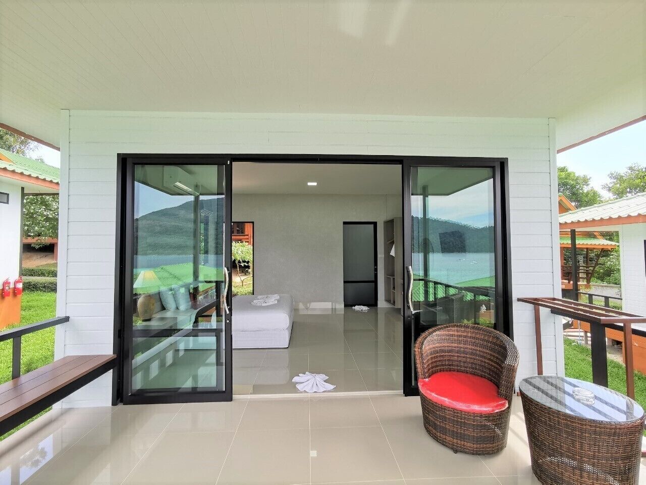 A modern hotel room with a glass door leading to a balcony, a wicker chair with a red cushion, and a small table.
