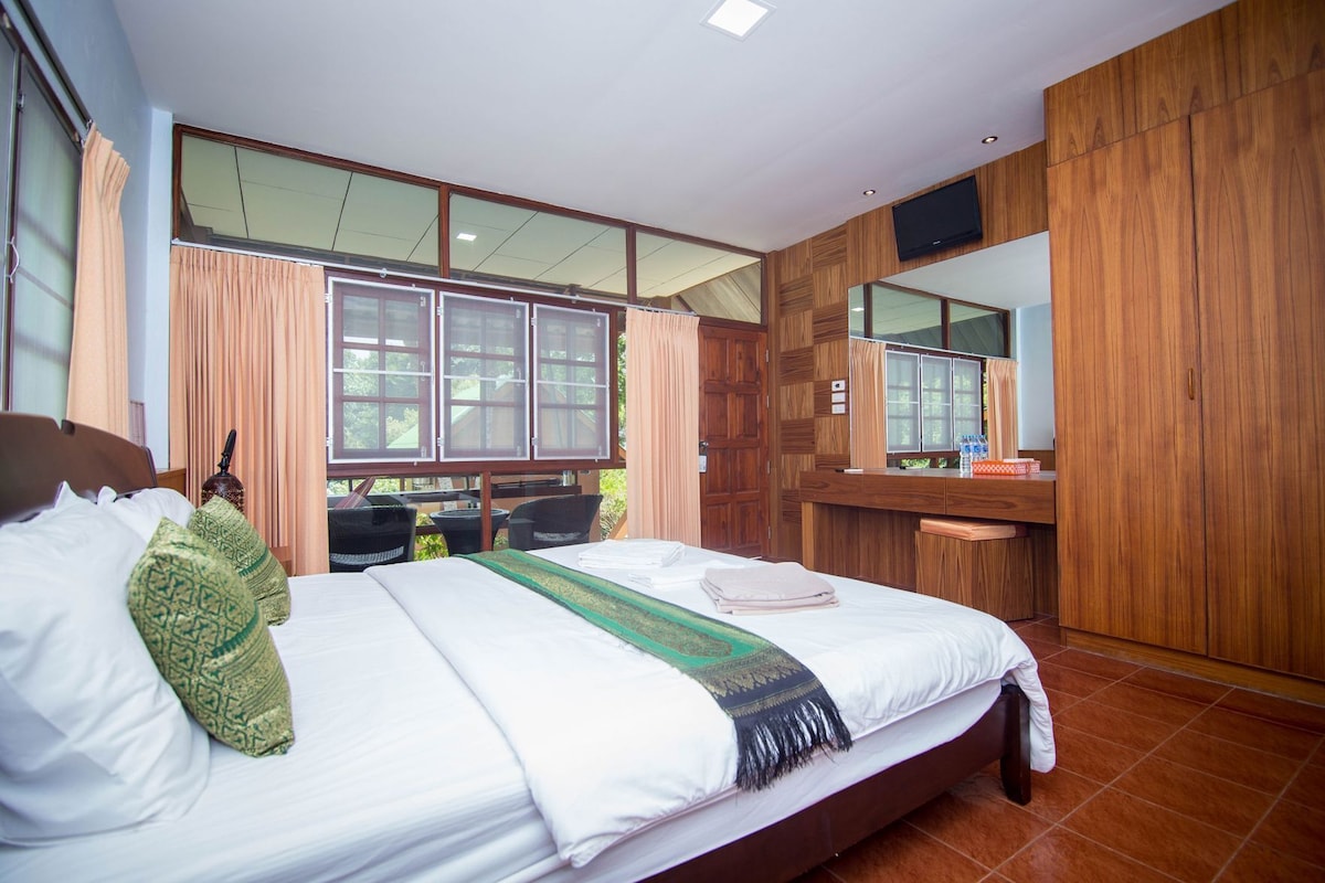 A hotel room with a large bed, a wooden wardrobe, a vanity with a mirror, and a window with curtains.