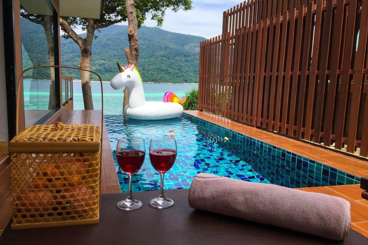 A poolside area with a wooden fence, a wicker basket, two glasses of red wine, and a rolled towel.