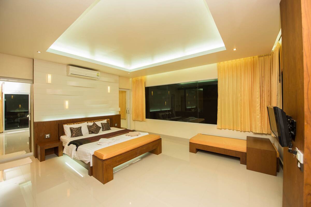 A modern bedroom with a large bed, a bench, a TV, and a large window with curtains.