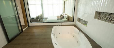 A modern bathroom with a large bathtub, a glass shower enclosure, and a view of a bedroom with a bed and curtains.