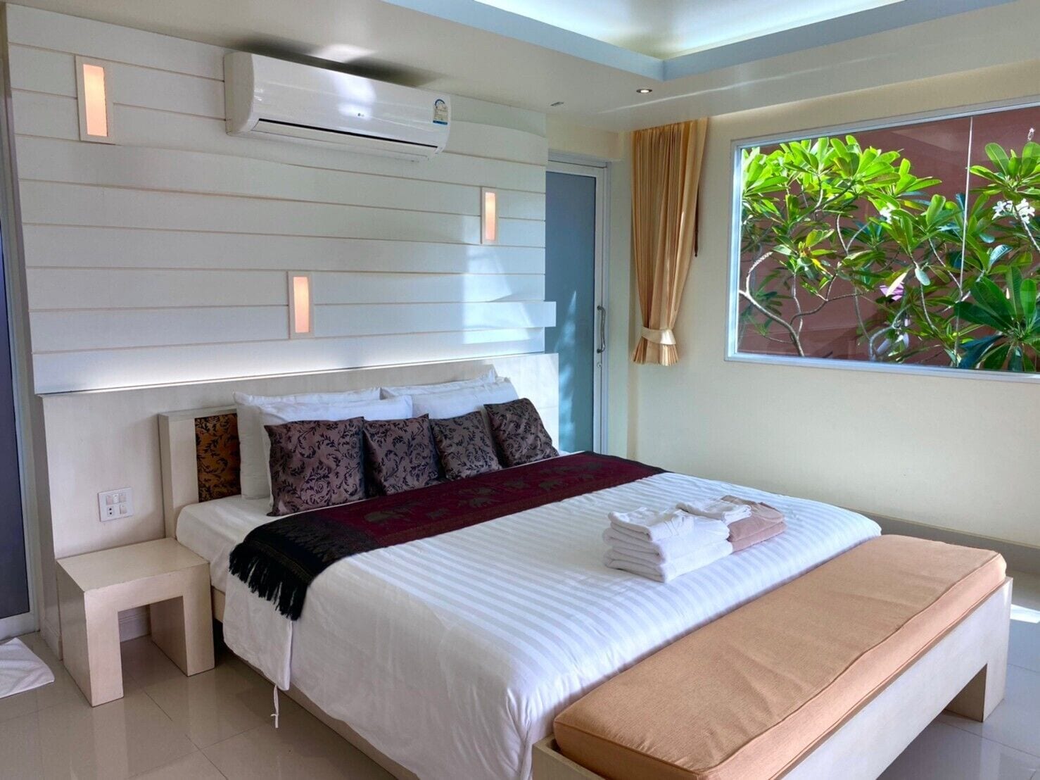 Ein modernes Hotelzimmer mit einem großen Bett, Nachttischen, einer Klimaanlage und einem Fenster mit Blick auf Grünflächen.