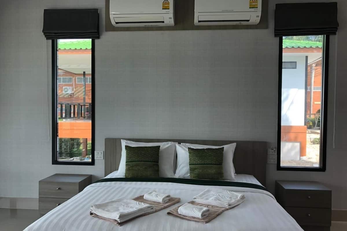 A hotel room with a large bed, two air conditioning units, a window with a view of buildings, and a nightstand with a lamp.