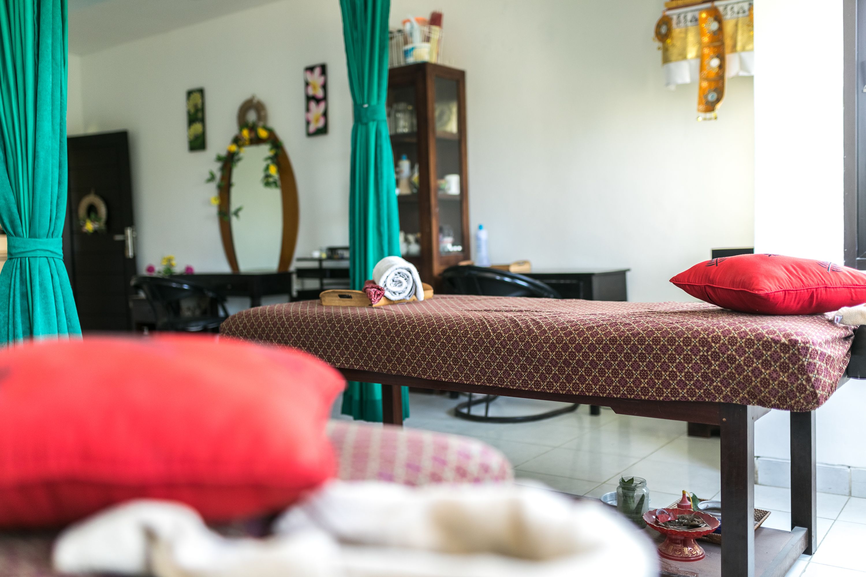 A massage table with red and white pillows, a wooden cabinet, and a mirror in a room with teal curtains.