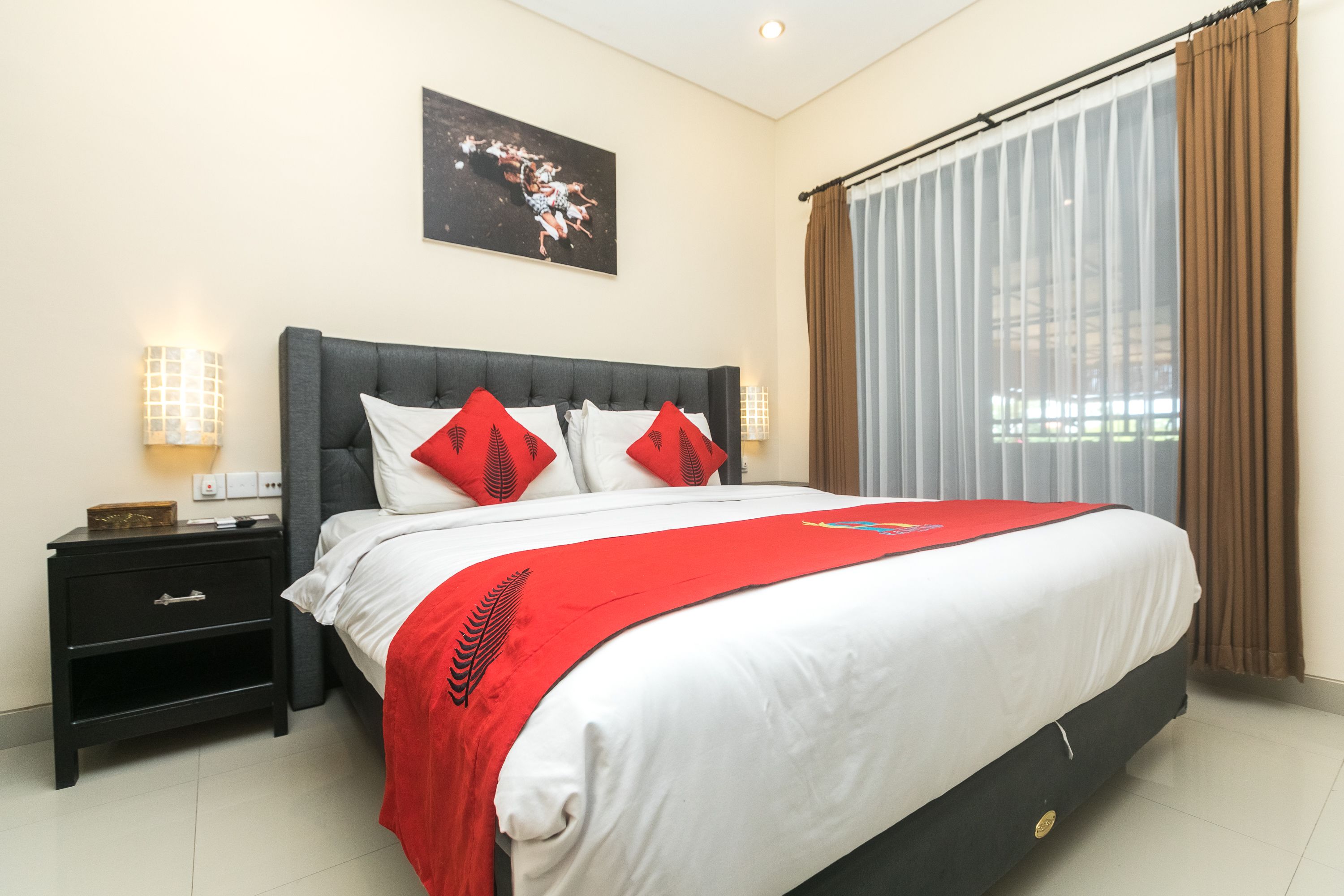 Een net opgemaakt bed met een wit-rode sprei, een donker hoofdbord en twee rode kussens. Er is een nachtkastje met een lamp en een klein tafeltje naast het bed. In de achtergrond is een groot raam met dunne gordijnen zichtbaar.