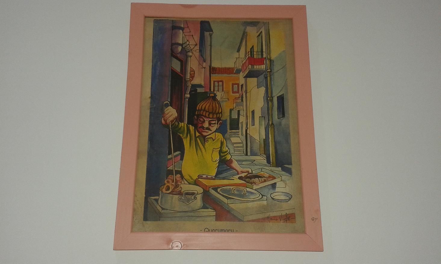 A framed artwork depicting a street scene with buildings and a person cooking.