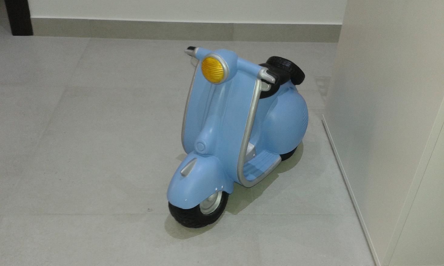 A blue scooter with a yellow headlight and black seat, parked indoors on a tiled floor.