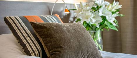 A neatly made bed with striped and solid pillows, a vase of white flowers, and a wall-mounted lamp.
