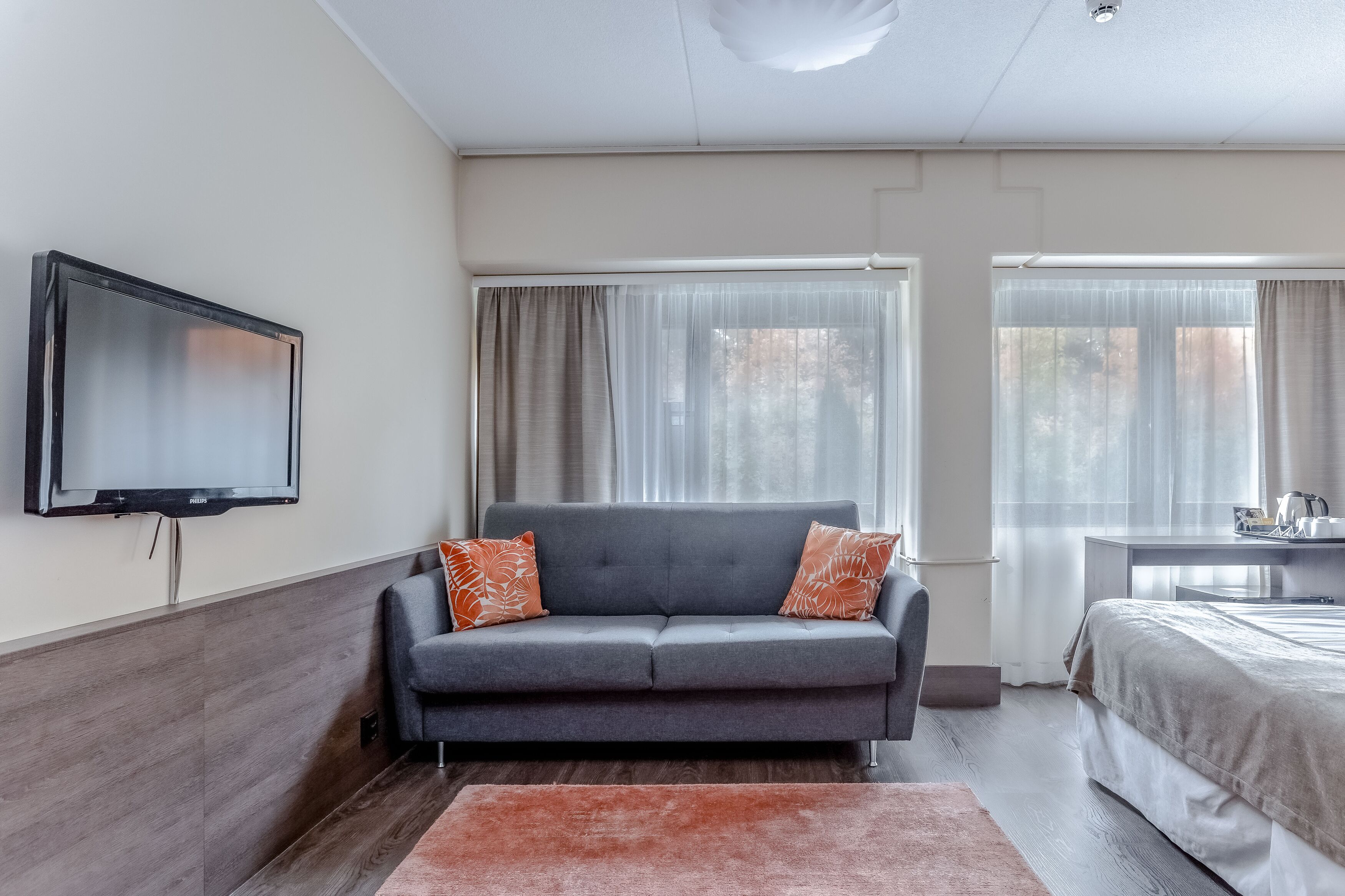 A modern hotel room with a grey sofa, a flat-screen TV, a bed, and a desk with a chair.