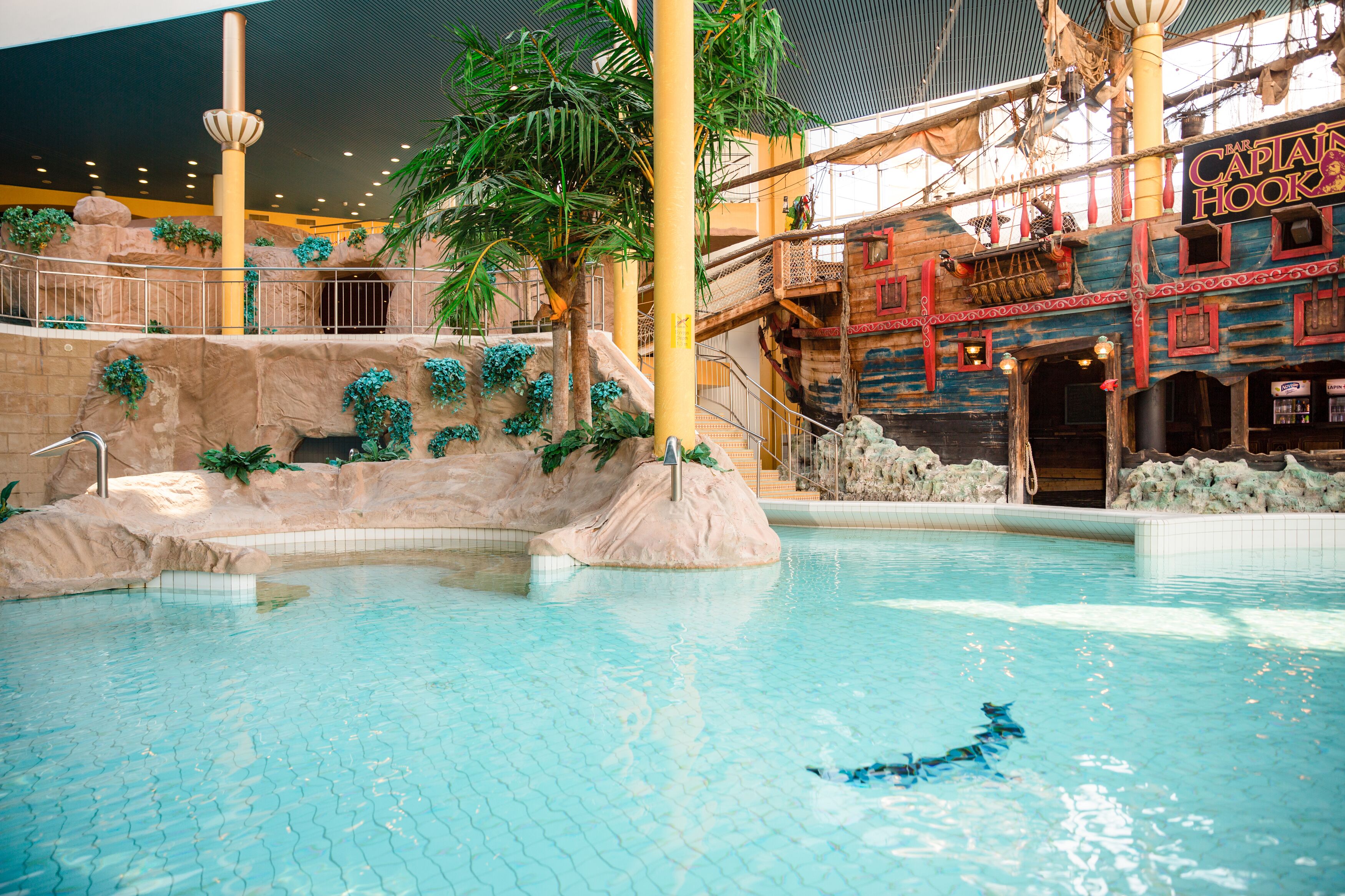 An indoor pool with a tropical theme, including a rock formation and palm trees.