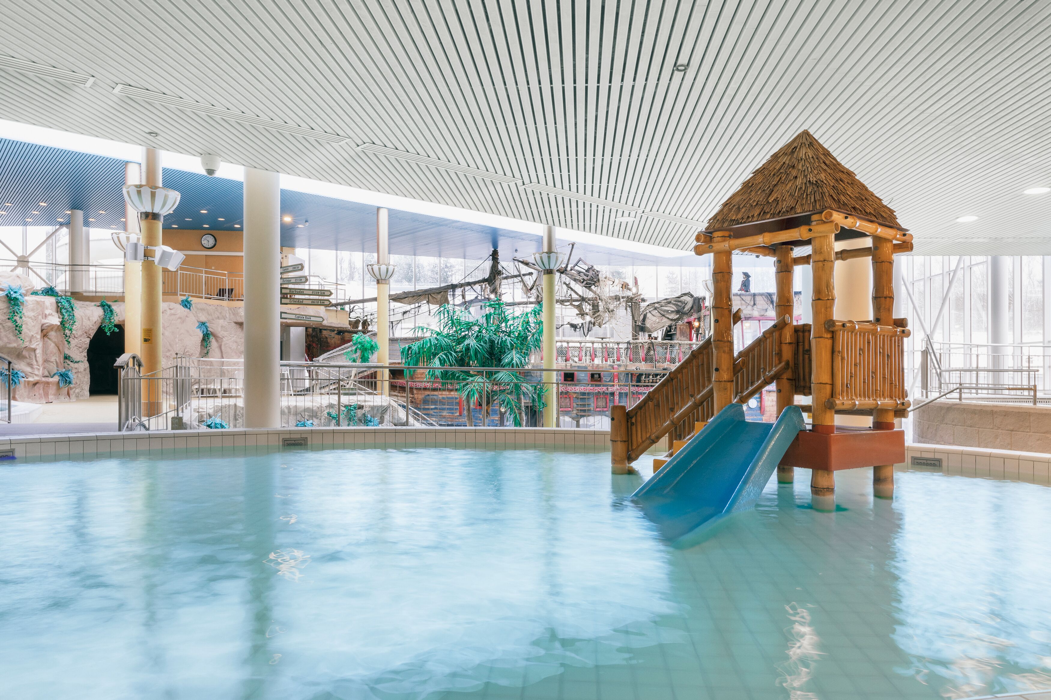 Parque acuático interior con una estructura de juego de madera, toboganes y una piscina grande.