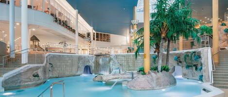 An indoor swimming pool with a waterfall feature, surrounded by rock formations and tropical plants.