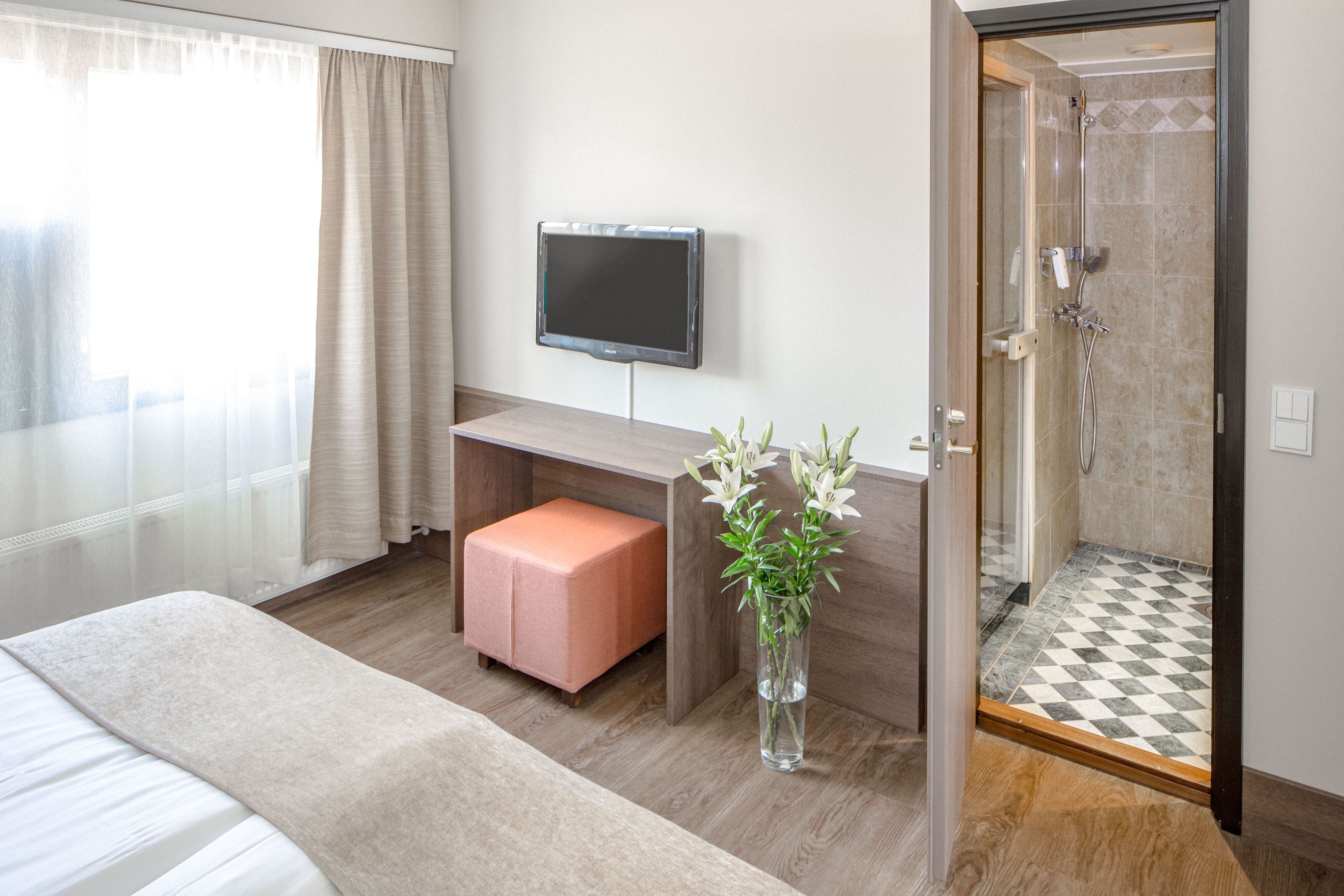 A hotel room with a bed, a desk with a pink ottoman, a TV mounted on the wall, a vase with flowers, and a shower area.